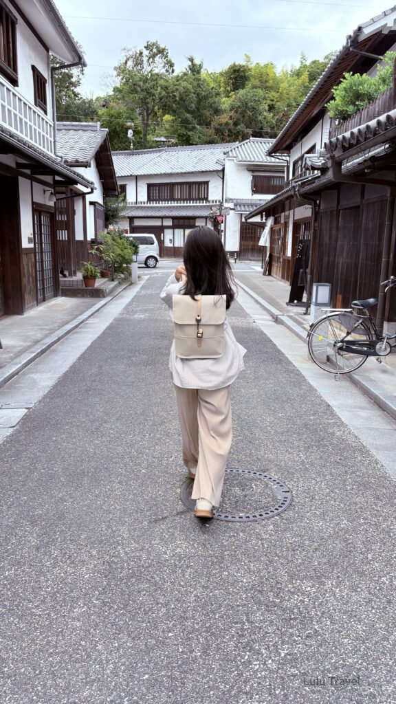 Slow Travel in Japan Kurashiki: An Afternoon Without an Agenda 日本傳統老街巷道中,一名旅人背著米色後背包行走在木造町屋之間,兩側是白牆深色木窗的日式建築與停放的腳踏車,街道安靜整潔,呈現慢步調的日本生活風景
