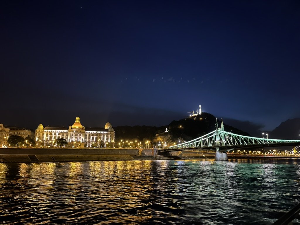 布達佩斯旅遊|第 1 次夜遊多瑙河 布達佩斯遊船景觀,遠方的自由橋(Liberty Bridge)與蓋勒特丘陵燈火倒映在波光粼粼的水面上。