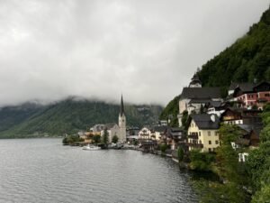 奧地利哈修塔特湖畔小鎮景色，彩色木屋沿著湖岸排列，教堂尖塔倒映在湖面上，遠方群山被雲霧環繞，呈現雨天的歐洲湖區童話風景