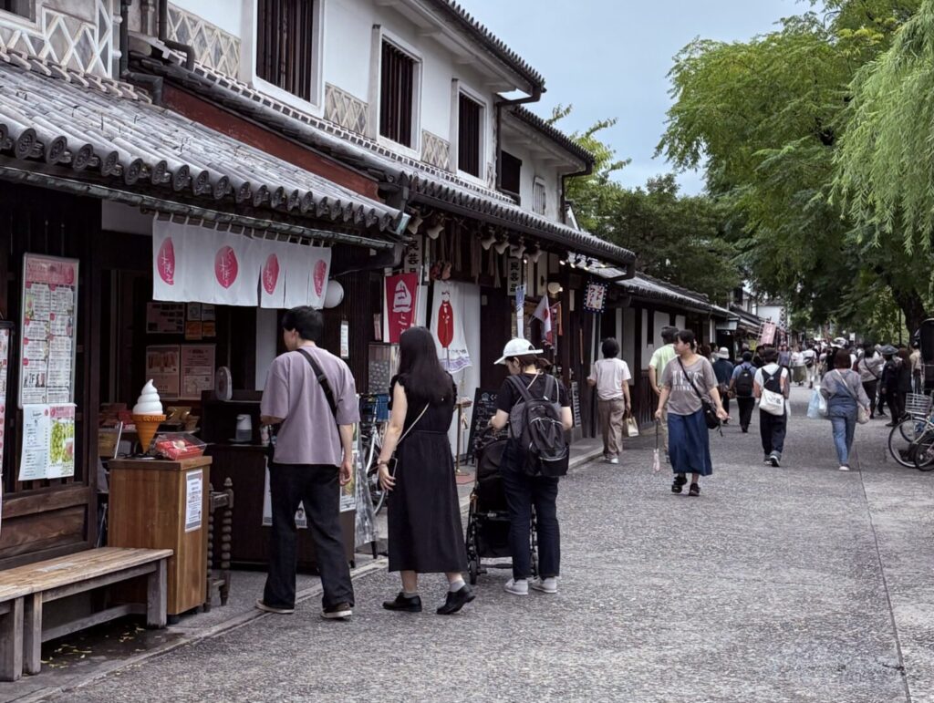 倉敷美觀地區|日本倉敷的 1 段散步旅遊記錄 日本岡山倉敷美觀地區,傳統町屋改建的商店街巷道,木造建築與綠意交錯,遊客沿著步道散步