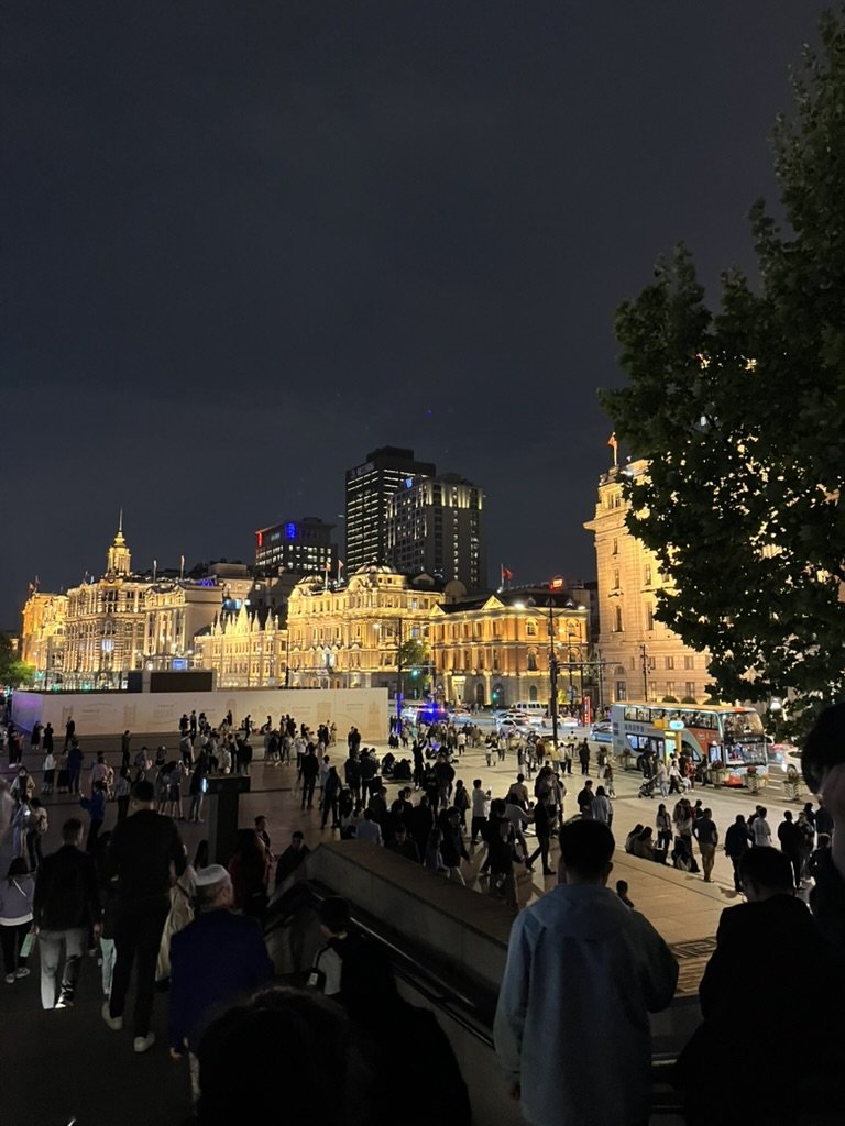 上海外灘夜景，人潮聚集於黃浦江畔，對岸為燈光點亮的歷史歐式建築群，展現上海夜晚的城市節奏與經典天際線