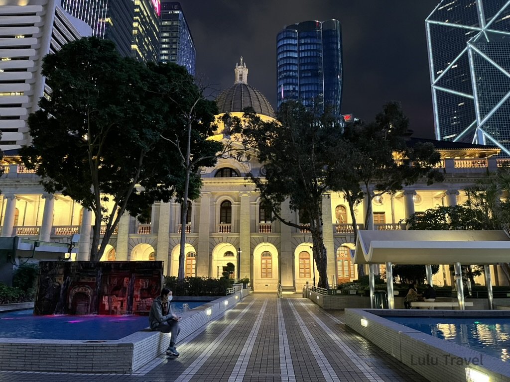 夜晚燈光下的歷史建築與現代高樓並列，前方廣場有人靜坐，展現城市中舊與新的對比景色