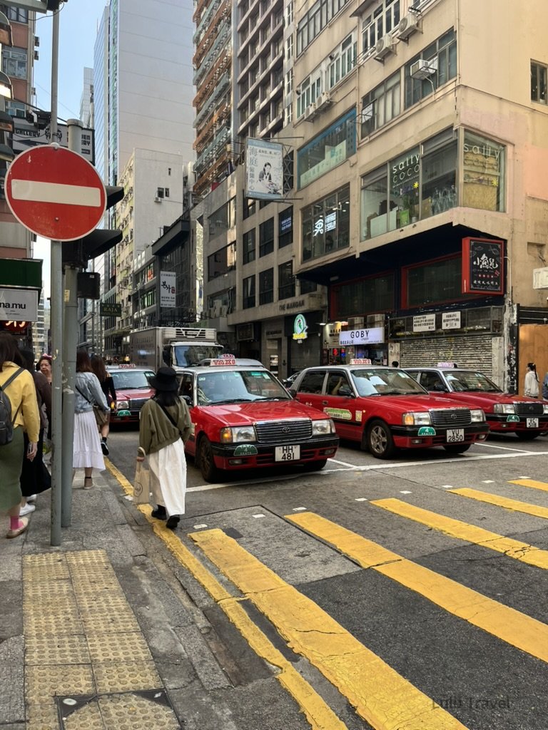 香港中環街景，紅色計程車行駛在狹窄街道上，兩側為高密度商業大樓與行人交錯的城市畫面