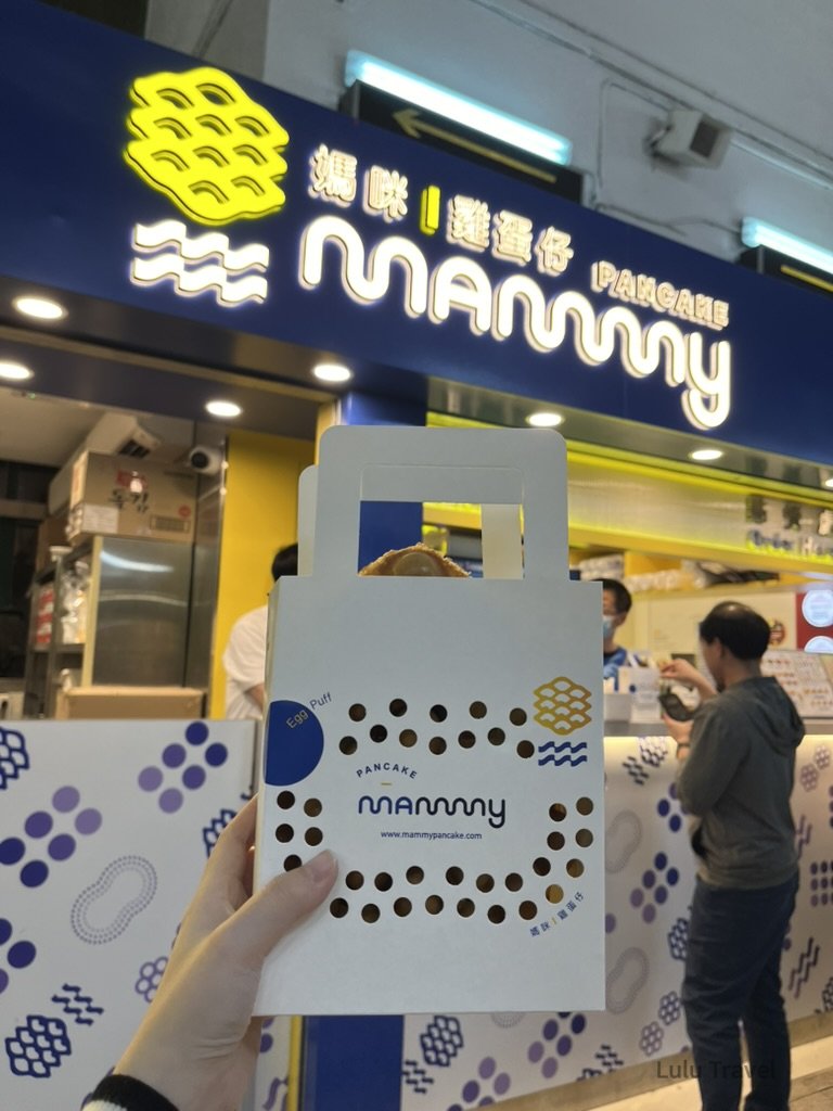 香港街頭 Mammy Pancake 雞蛋仔店，旅人手持外帶包裝站在店前，體驗香港經典街頭小吃