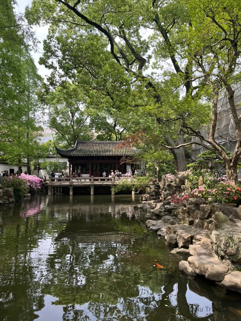 上海豫園園景，傳統江南園林建築臨水而建，池塘、假山與綠樹環繞，遊客於園中漫步