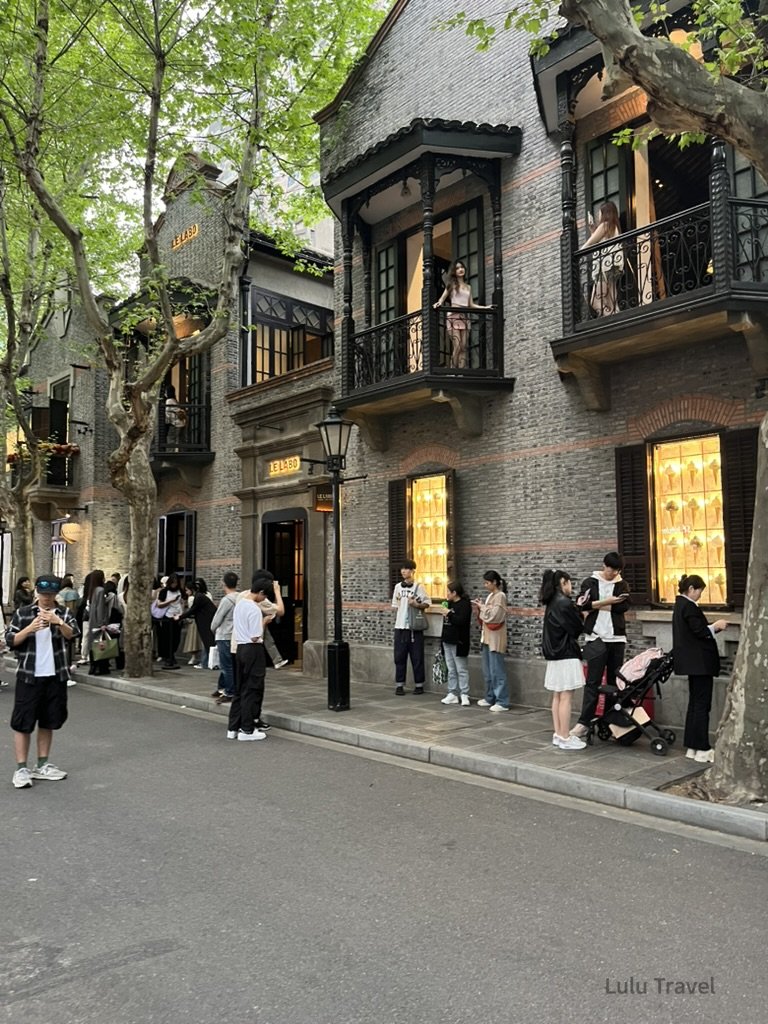 上海街頭林蔭大道旁的歷史建築外觀，灰磚洋樓立面與陽台設計，行人聚集拍照與排隊，呈現上海租界風格街景