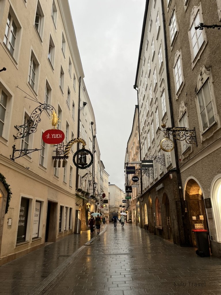 奧地利薩爾斯堡老城街道，雨後石板路與歷史招牌，行人漫步其間