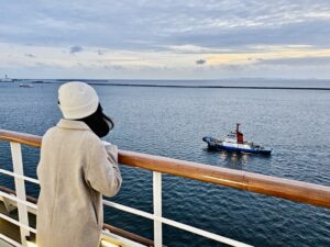 日本郵輪旅行 | 高雄出發歌詩達莎倫娜號的 10 個真實心得 歌詩達莎倫娜號郵輪甲板看海實況,旅客穿著大衣與毛帽倚靠欄杆,欣賞海面上的引水船與風景,真實呈現海上航行時的氣溫與防風保暖穿搭。