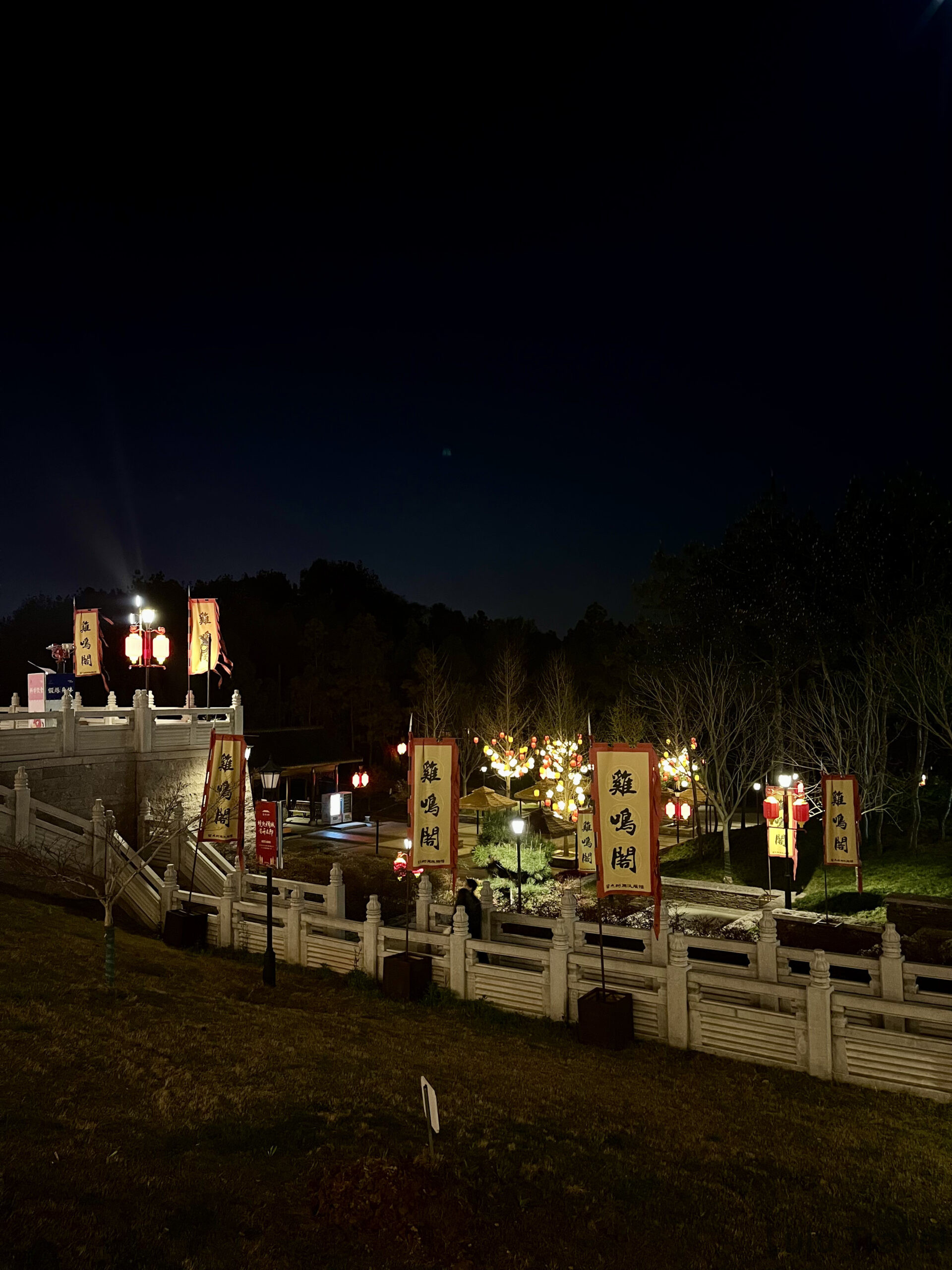 義烏雞鳴閣公園入口夜景，白色石欄杆旁排列多面寫有「雞鳴閣」的紅黃旗幟，背景為彩色燈籠裝飾的樹叢與夜空