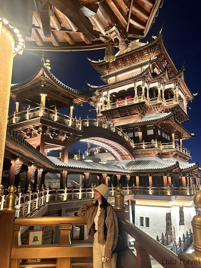 在義烏雞鳴閣夜景前拍照留念，戴帽女生站在木構迴廊旁仰望多層傳統古塔，暖色燈光映照整座建築與深藍夜空