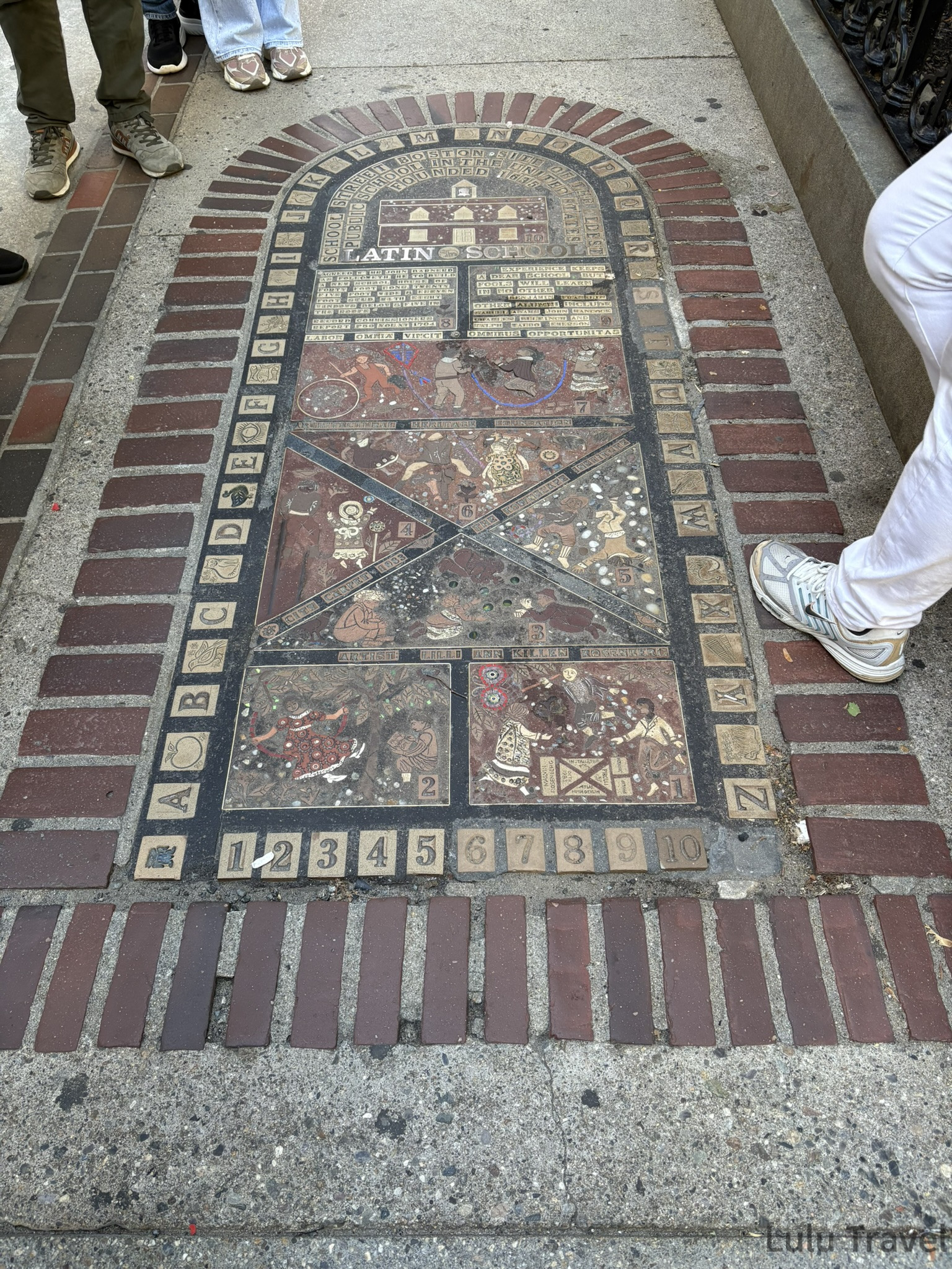 波士頓自由之路沿途的 Boston Latin School 紀念地磚，鑲嵌於人行道上，以磚紅色框架圍繞，內部有字母、數字與歷史場景的馬賽克圖案，記錄美國最古老公立學校的創立歷史。