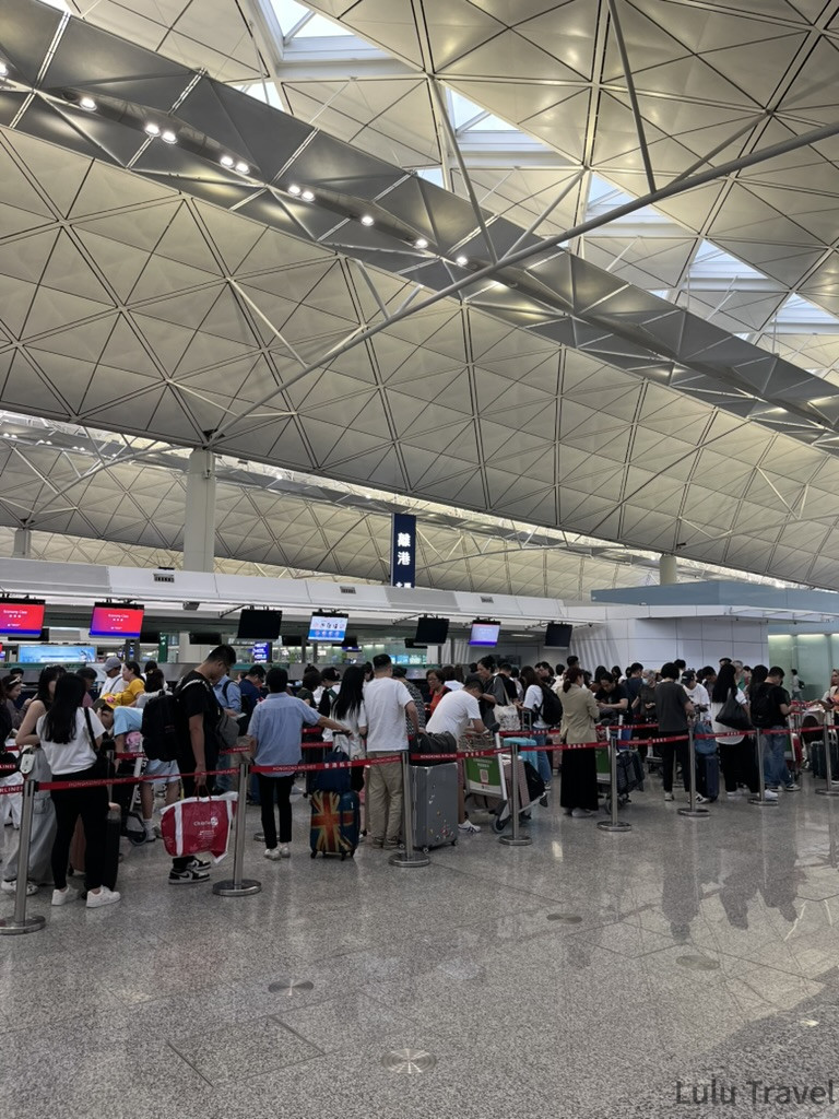 香港機場 check-in 大廳，旅客拉著行李在香港航空等多家航空的報到櫃台前排隊