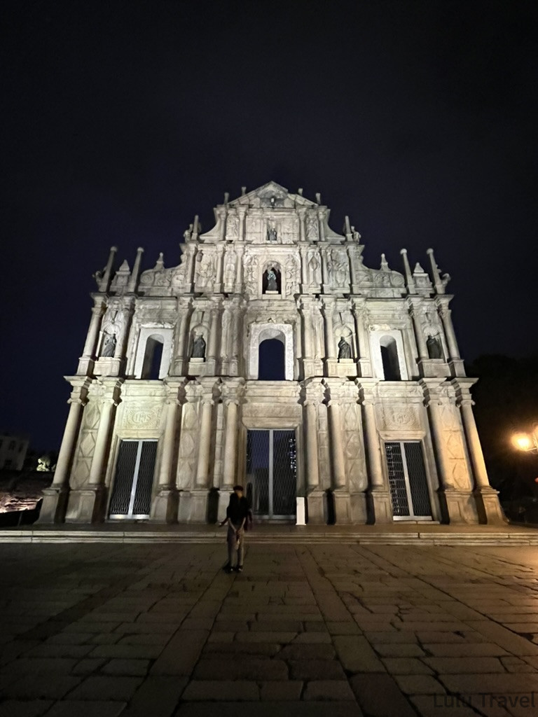 澳門大三巴夜景全貌，燈光照亮巴洛克風格石砌立面，石階上有遊客拍照留影