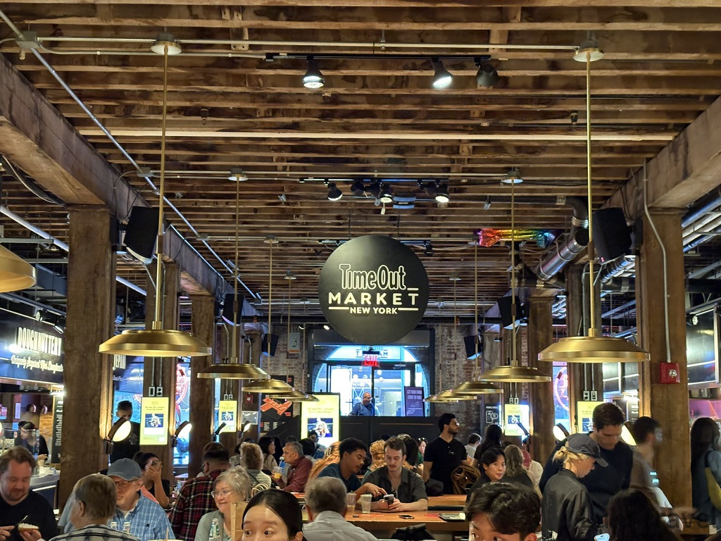 紐約 DUMBO 區 Time Out Market 工業風室內用餐區，木質結構搭配工業吊燈與黑色圓形標誌，是 J 人行程中理想的食物中繼站。