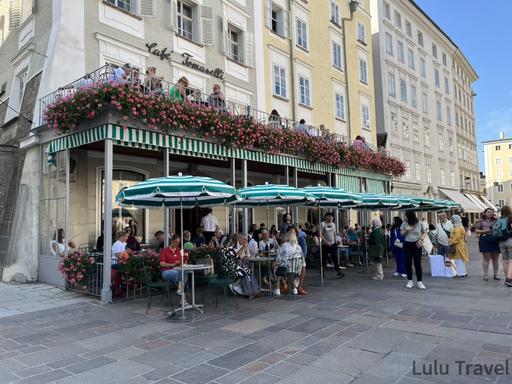 薩爾斯堡景點 Café Tomaselli 外觀，綠白條紋遮陽棚下坐滿旅客，二樓陽台盛開粉色花卉