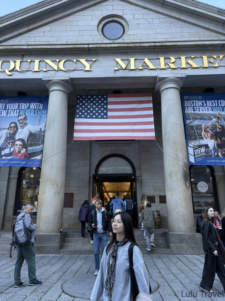 昆西市場（Quincy Market）正面外觀宏偉的希臘復興式石造建築入口，上方刻有「QUINCY MARKET」金色字樣。巨大的石柱間掛著一面美國國旗與兩幅藍色宣傳海報。