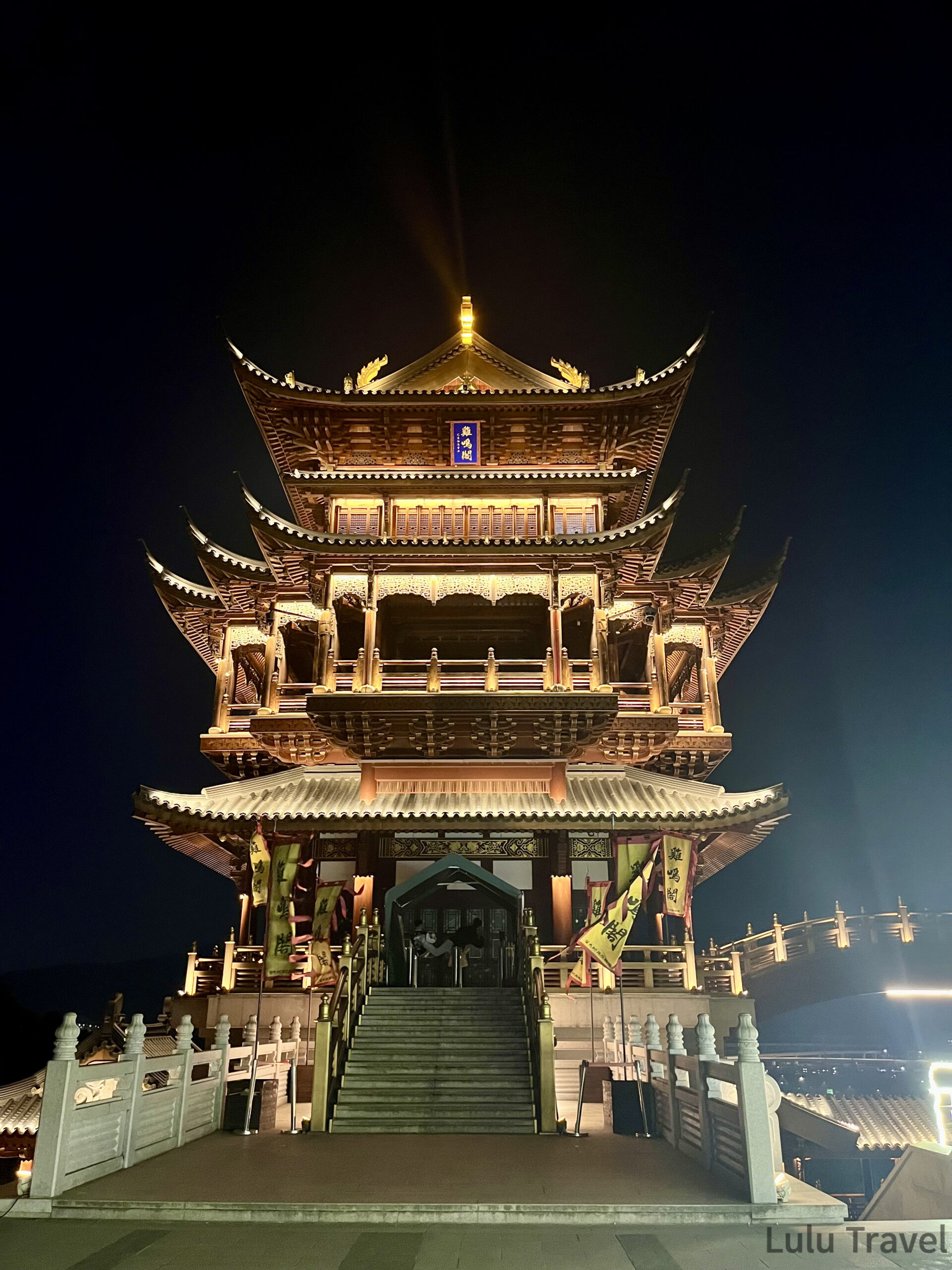 義烏雞鳴閣正面夜景，五層中式木構古塔正對鏡頭，頂部金色燈光射向夜空，入口兩側豎立彩旗，石階通往大門