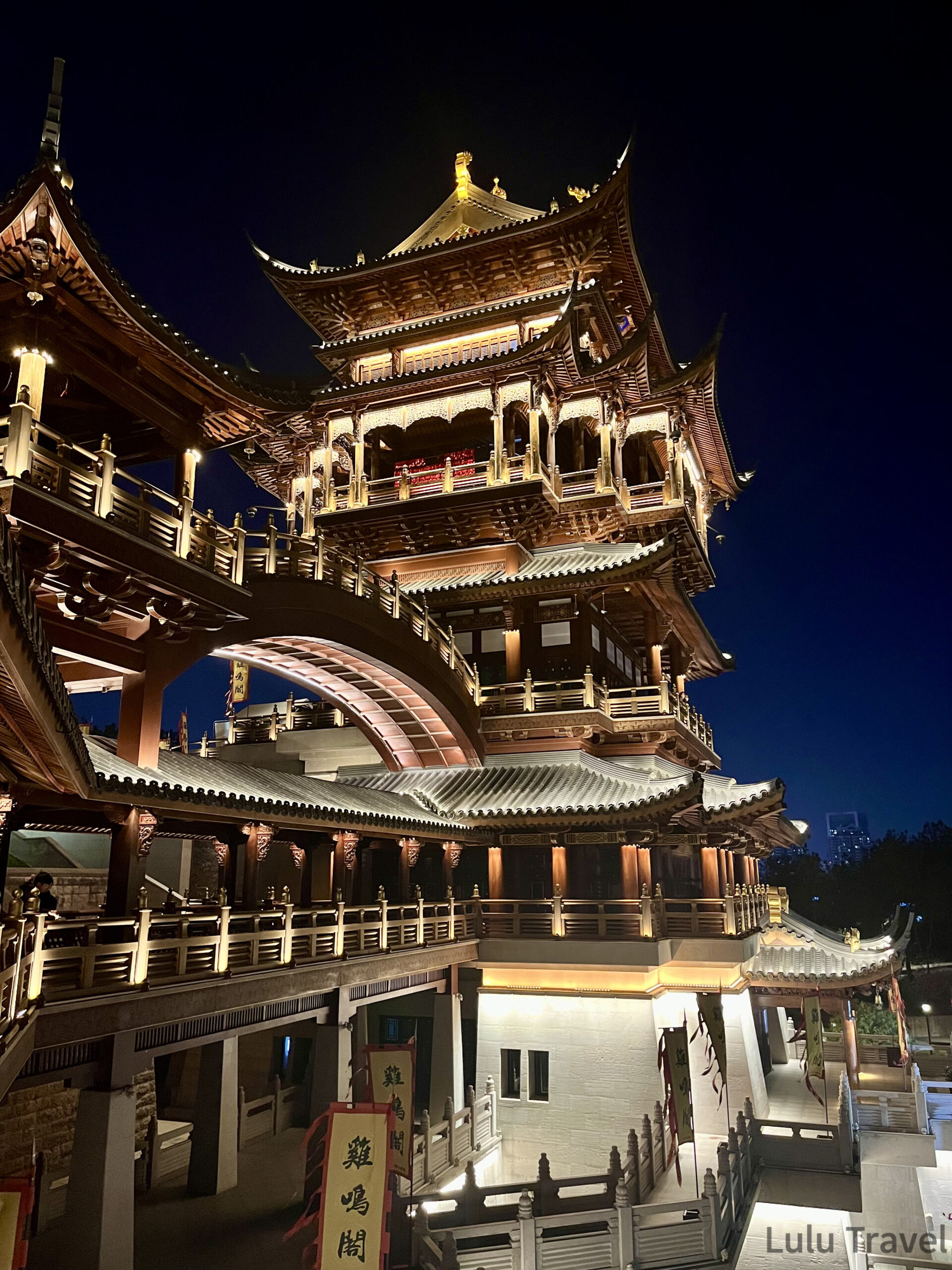 義烏雞鳴閣多層樓閣夜景全貌，傳統木雕飛簷層層疊起，暖黃燈光打亮整座建築，前景可見雞鳴閣招牌旗幟