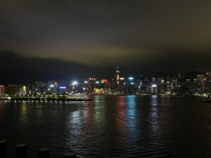 香港機場驚魂|違禁品被扣留、錯過 check-in 多待 1 晚 一張在夜間拍下的香港天際線照片。遠處的摩天大樓在夜色中發出閃爍的光芒,維多利亞港的水面也倒映著這些燈光。天空陰雲密布,右側隱約可見山脈。左側水面上有一艘渡輪,渡輪上的燈光格外明亮。