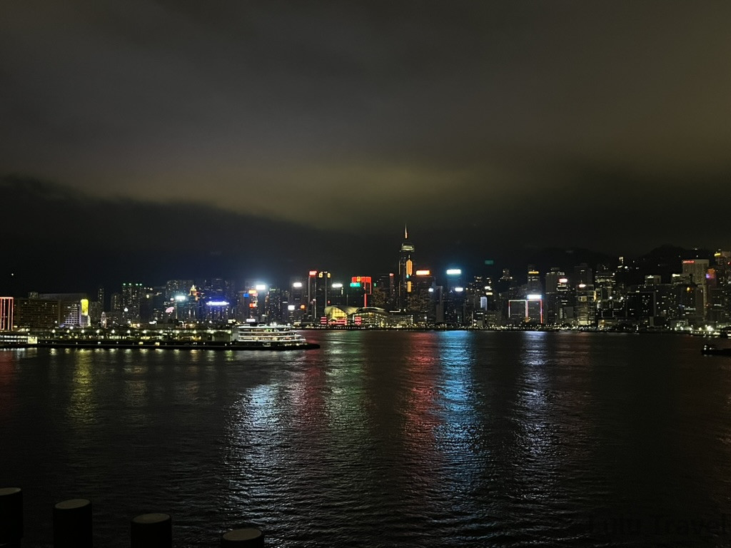 一張在夜間拍下的香港天際線照片。遠處的摩天大樓在夜色中發出閃爍的光芒，維多利亞港的水面也倒映著這些燈光。天空陰雲密布，右側隱約可見山脈。左側水面上有一艘渡輪，渡輪上的燈光格外明亮。