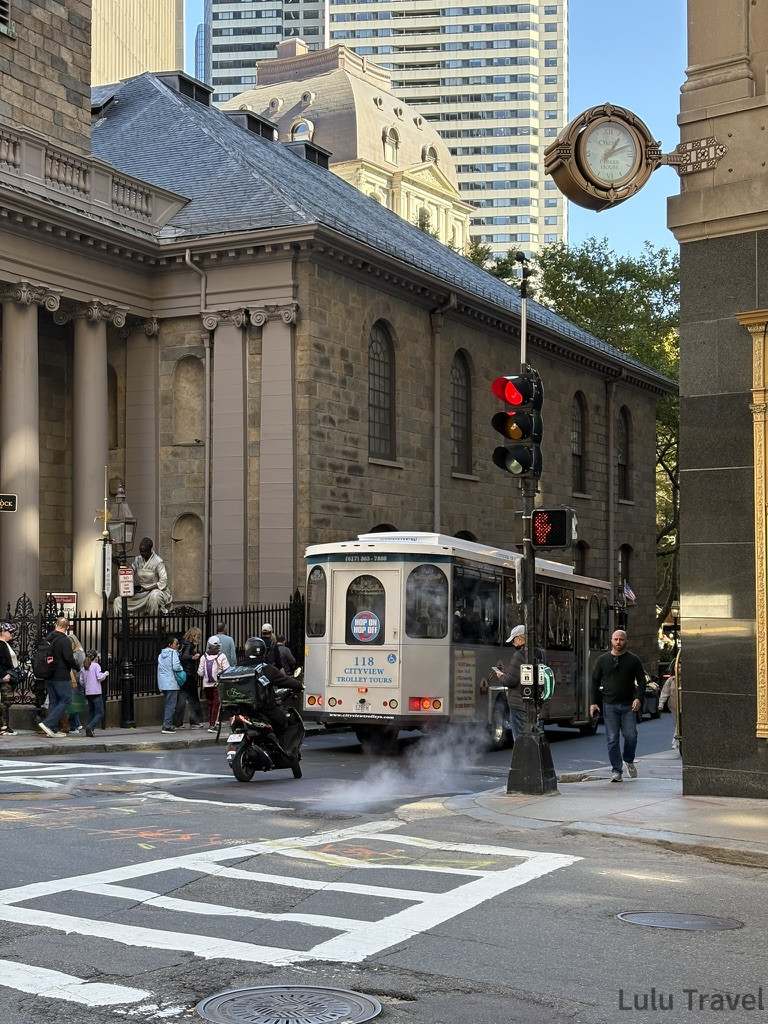 國王禮拜堂與街景是充滿歷史感的古老石造教堂（King's Chapel）與現代高樓並存。畫面中有一輛復古觀光電車（Trolley）正經過十字路口，展現波士頓新舊交融的城市日常。
