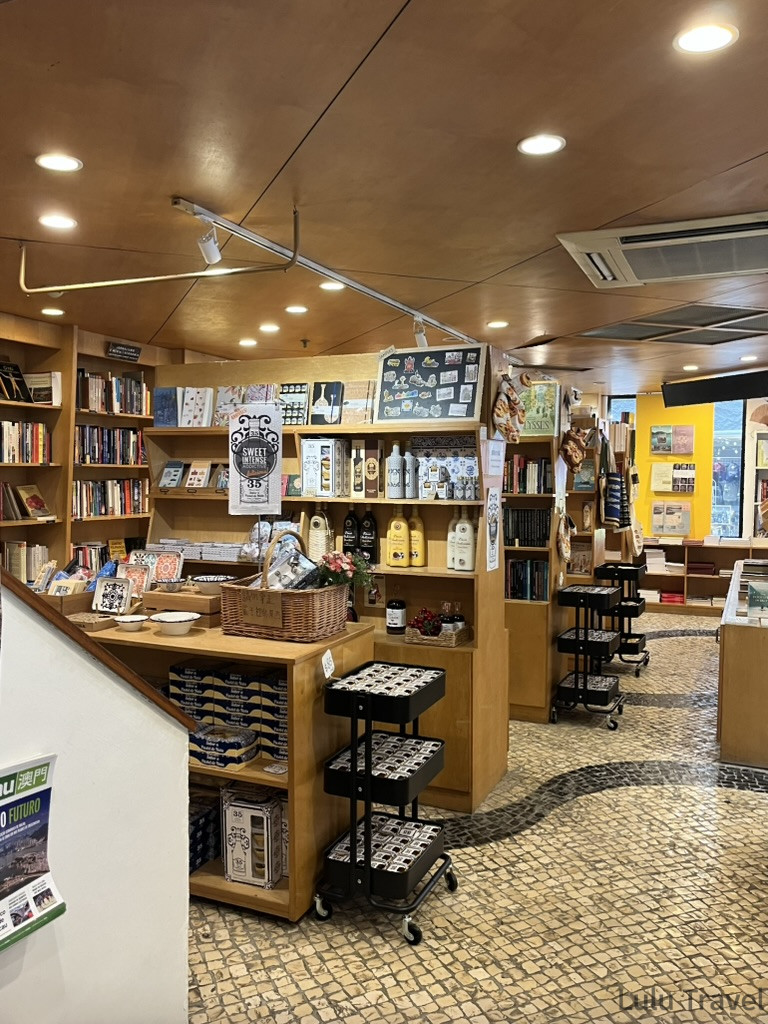 澳門葡文書局內部，木質書架陳列書籍與葡式食品，石板地面延伸至採光良好的書局深處
