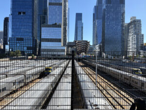 紐約 Hudson Yards 景觀實拍，從高處俯瞰密集的 LIRR 鐵道與停靠的銀色列車，背景為著名的蜂巢狀建築 The Vessel 與曼哈頓現代摩天大樓群，展現極致的都市工業尺度。