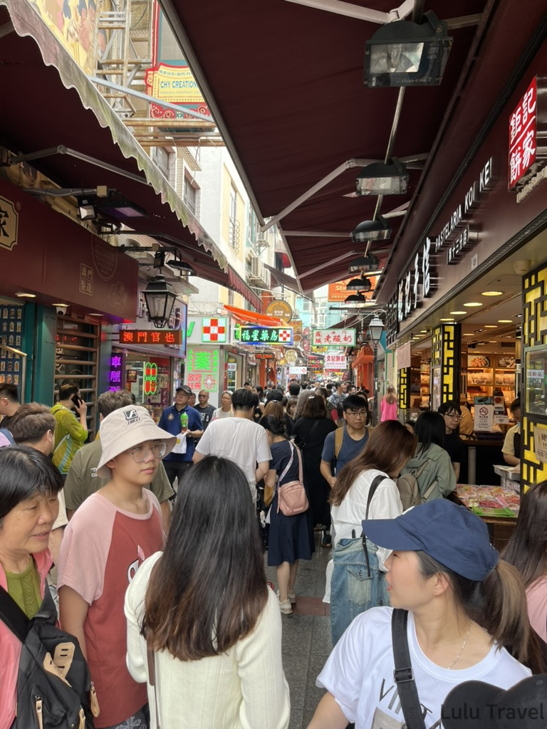 澳門官也街白天人潮，狹窄小吃街兩側密集排列各式澳門伴手禮攤位，旅客摩肩接踵