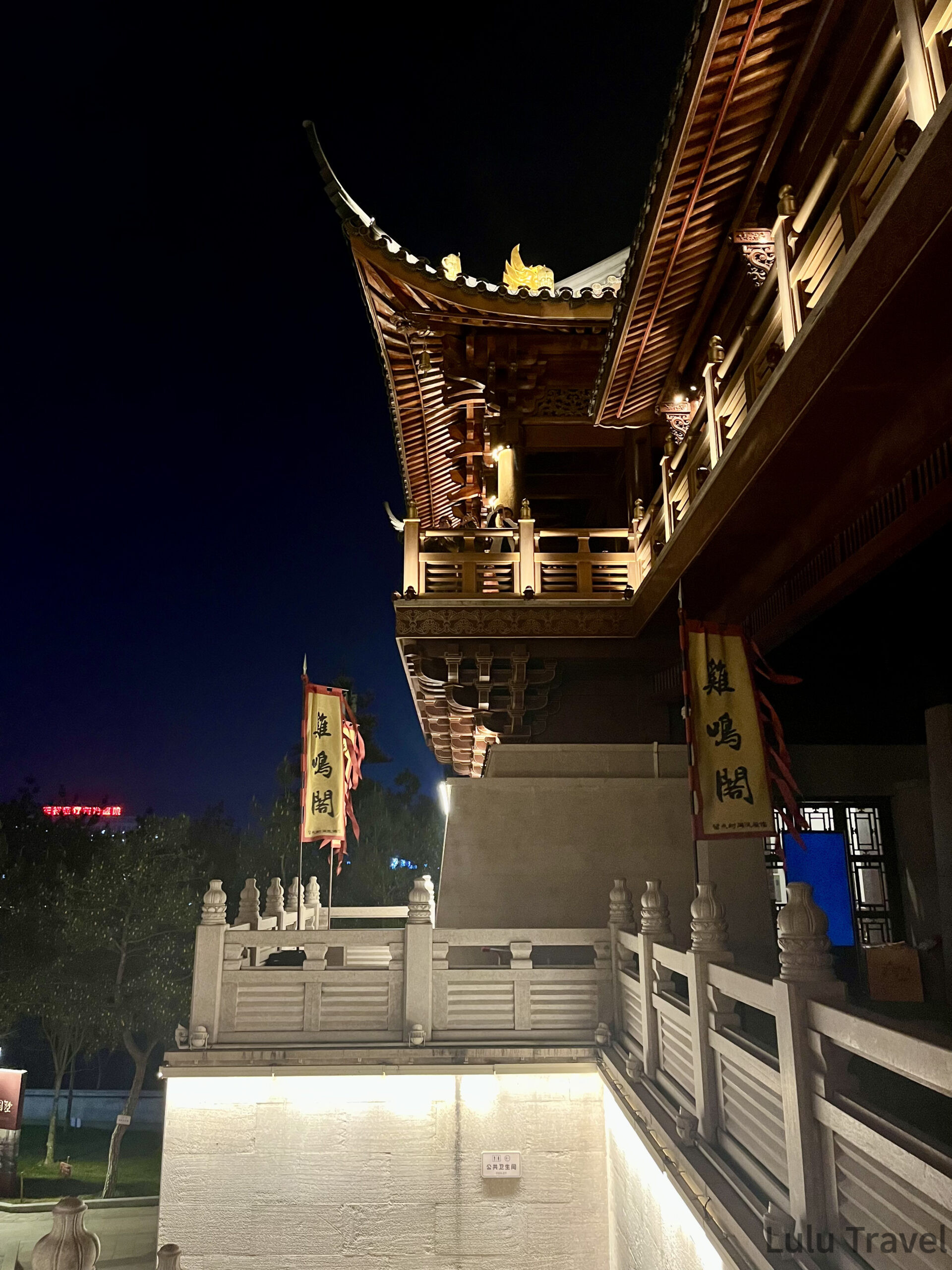 義烏雞鳴閣近景仰拍，中式飛簷屋頂與木構迴廊在夜燈下顯現精細雕刻，前景為白色石欄與雞鳴閣旗幟