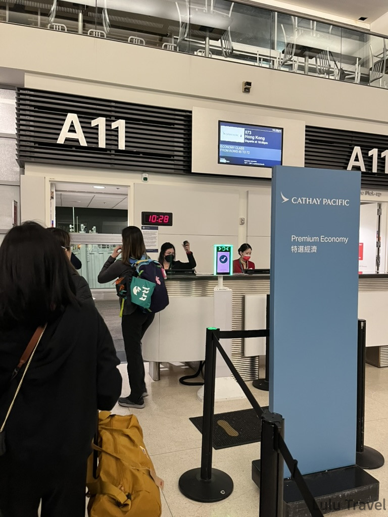 國泰航空（Cathay Pacific）的辦公櫃檯，兩名工作人員正在忙碌。櫃檯旁邊立著一塊藍色的牌子，上面寫著「Premium Economy 特選經濟」。在他們前方，幾位旅客正在排隊等候登機。