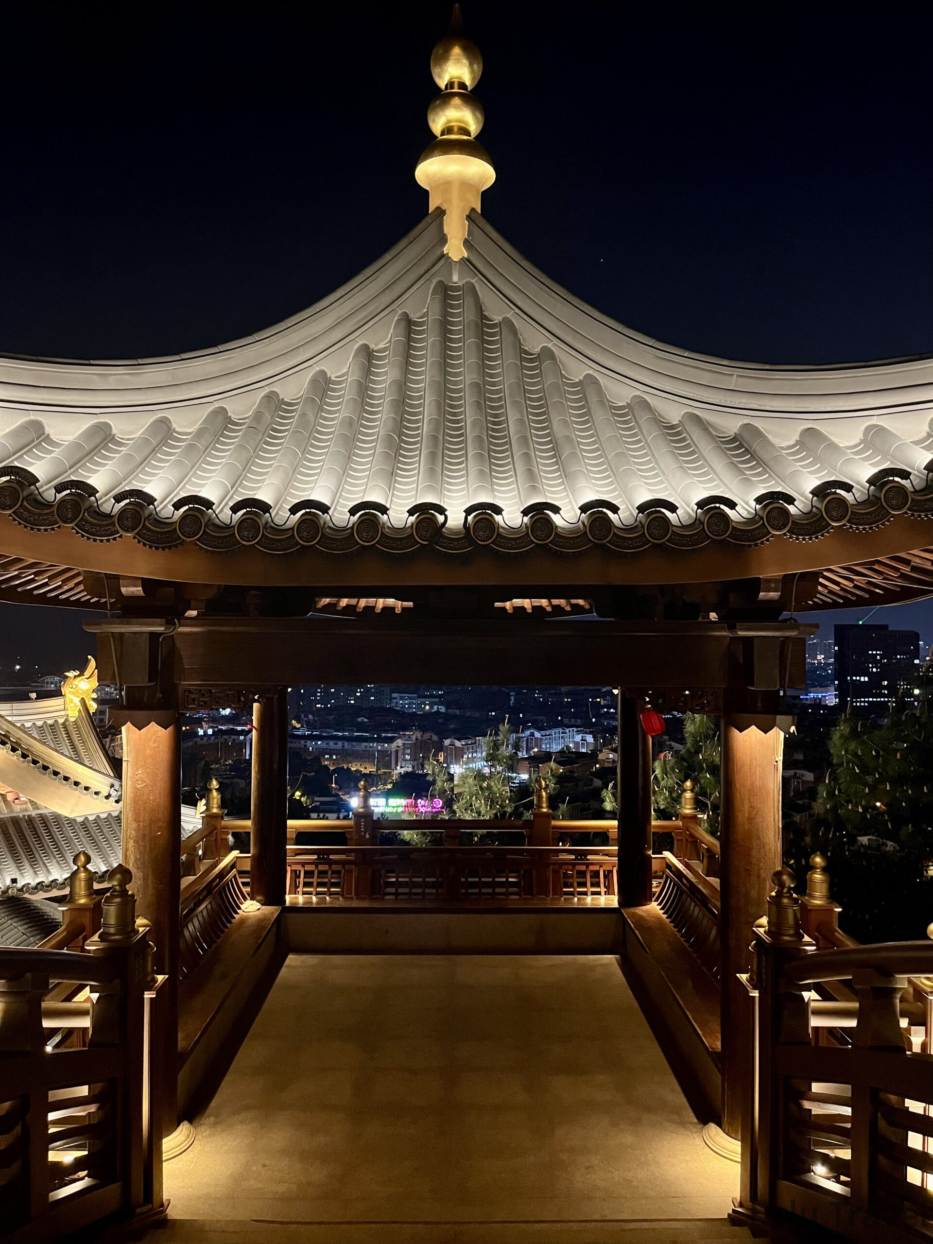 從義烏雞鳴閣涼亭內向外看的夜景，白色琉璃瓦亭頂與金色頂珠框住遠方義烏市區的夜間燈火