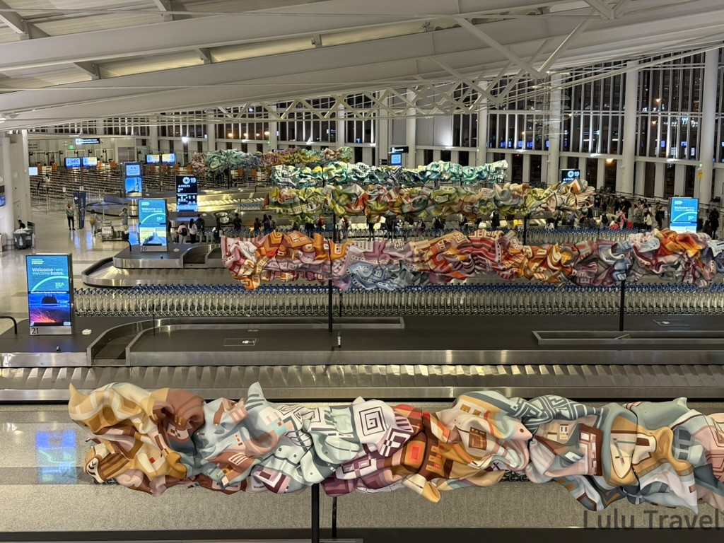 西雅圖機場（SEA-TAC）行李領取大廳實景，行李轉盤上方有色彩鮮艷的藝術裝置。