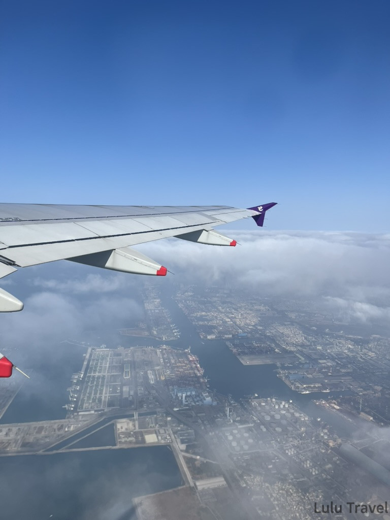 從飛機窗戶俯瞰的風景。畫面左上方可見機翼，翼尖帶有紫色香港快運標誌及紅色的平衡器。下方是廣闊的港口與工業區，可以看到筆直的水道、密集的貨櫃碼頭以及整齊排列的工廠建築，遠處則延伸至大海。