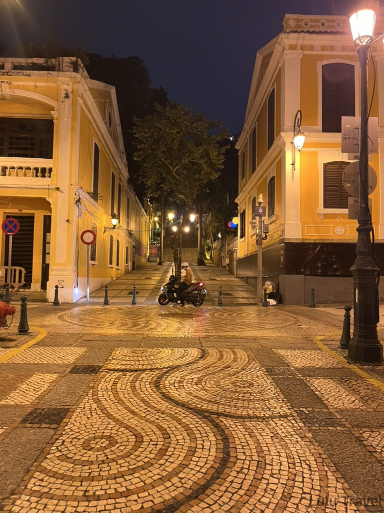 澳門半島夜晚的葡式圓石廣場，波浪紋石板地面反射路燈光芒，兩側為黃色葡式建築