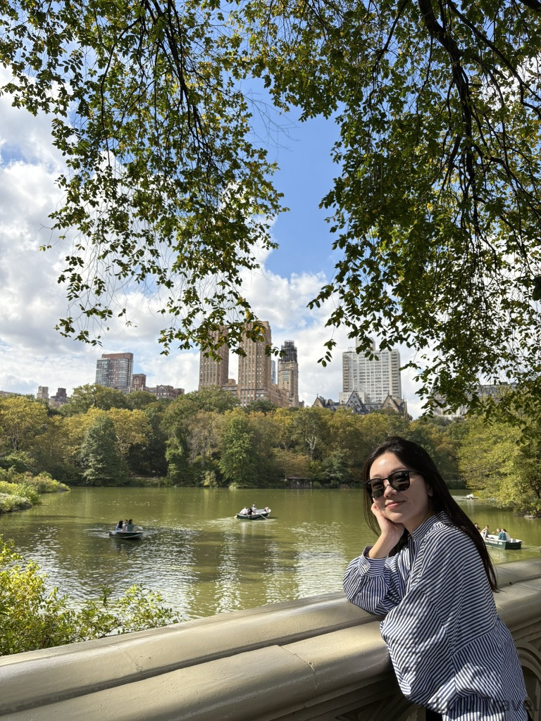 中央公園湖泊景觀，旅客倚靠欄杆欣賞對岸的聖雷莫公寓（The San Remo）雙子塔建築，湖面上有人正在划船，氣氛安靜從容。