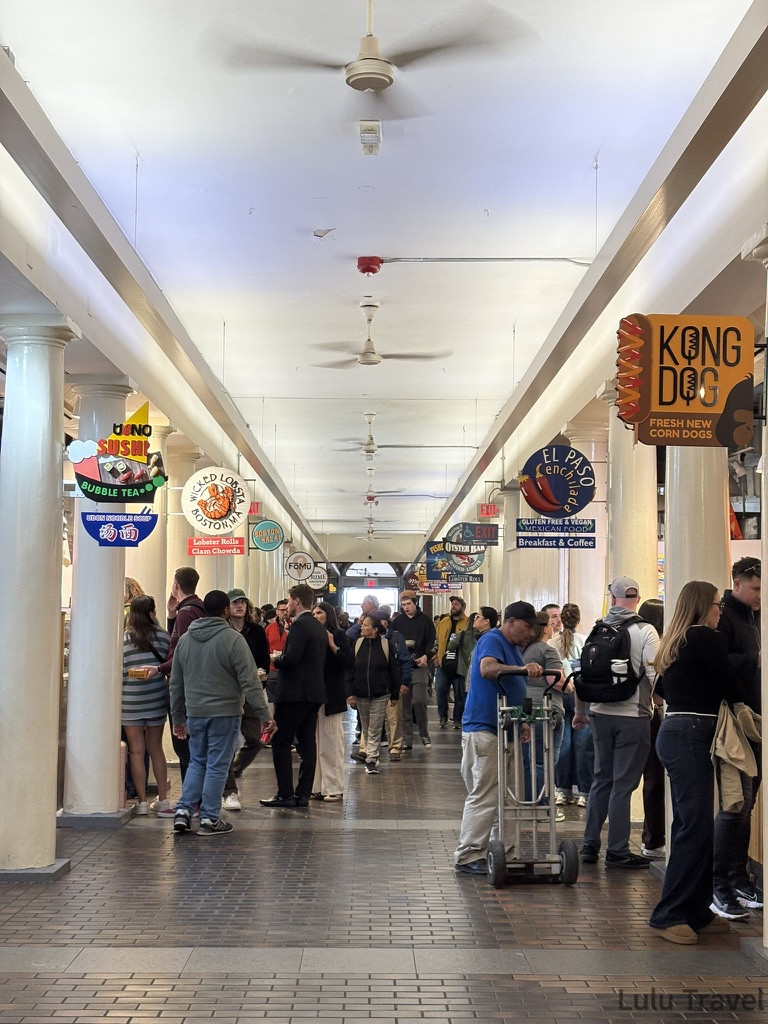 昆西市場 (Quincy Market) 內部熱鬧的市場長廊，天花板掛著吊扇，兩旁林立著各式餐飲招牌（如龍蝦堡、韓式熱狗、壽司等），遊客穿梭其中尋找美食。